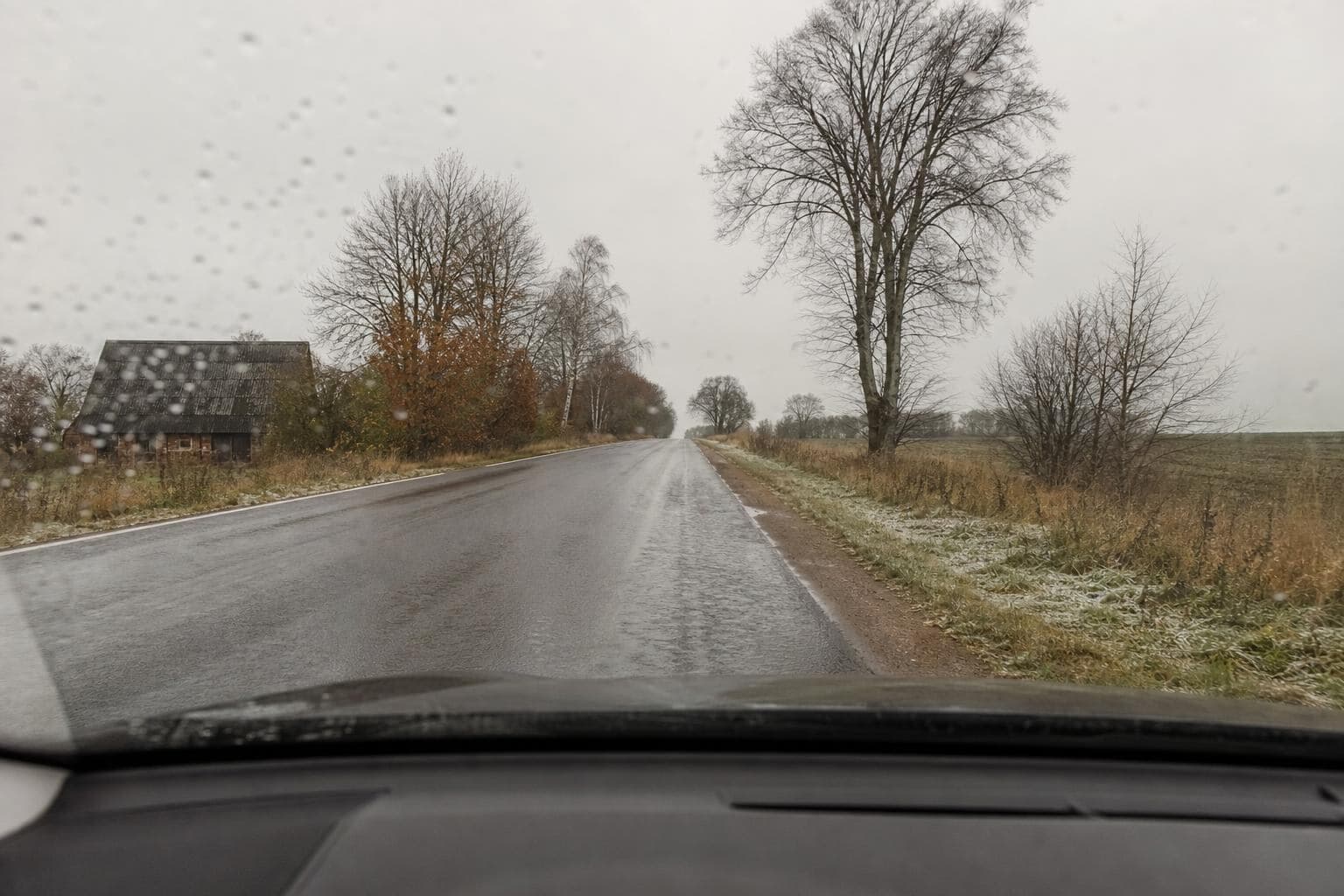 Våd landevej set fra førersædet gennem en forrude med regndråber, nøgne træer og rim på græsset en grå dag