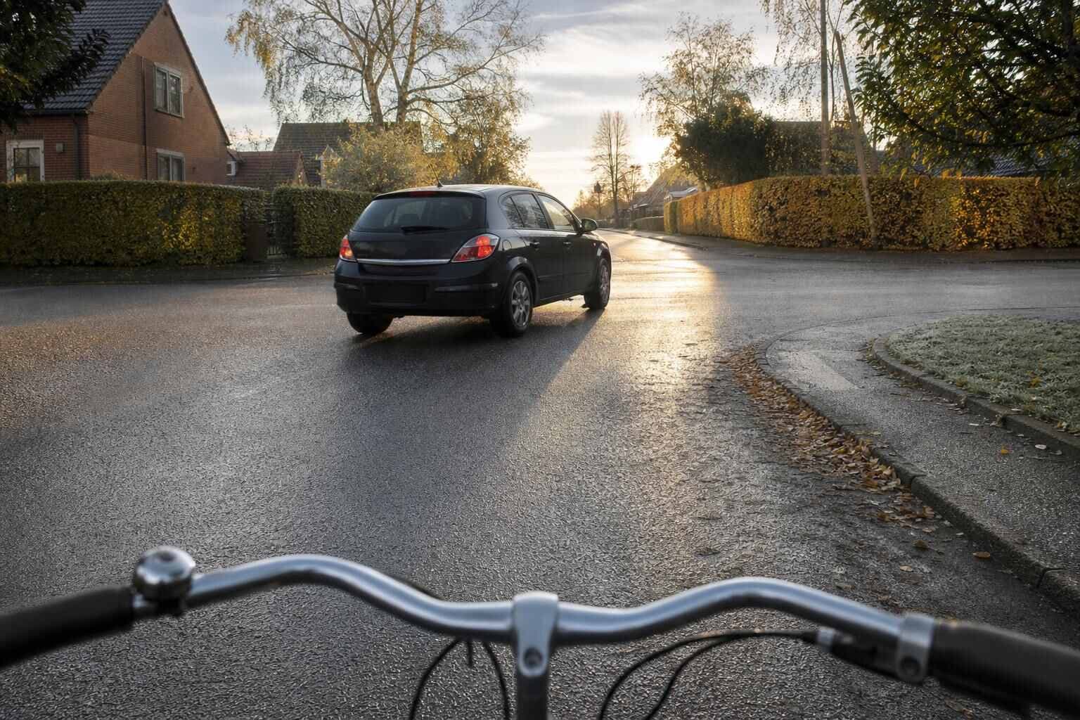 Cykelstyr set bagfra ved et lille villakryds, hvor en mørk bil svinger foran i lavt efterårslys på våd asfalt