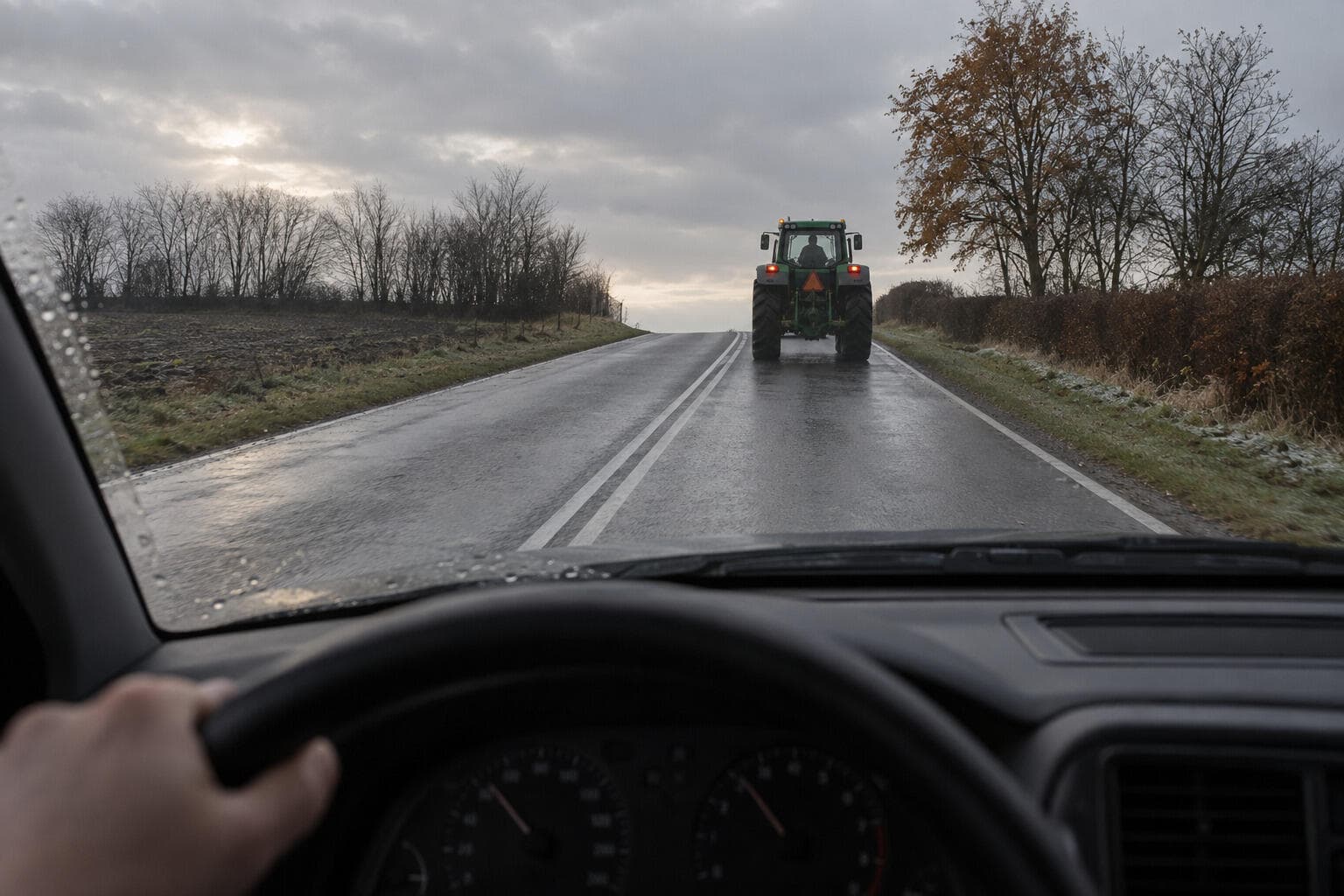 Landevej set fra førersædet mod en bakketop med dobbelt ubrudt hvid midterlinje, en grøn traktor foran og våd asfalt